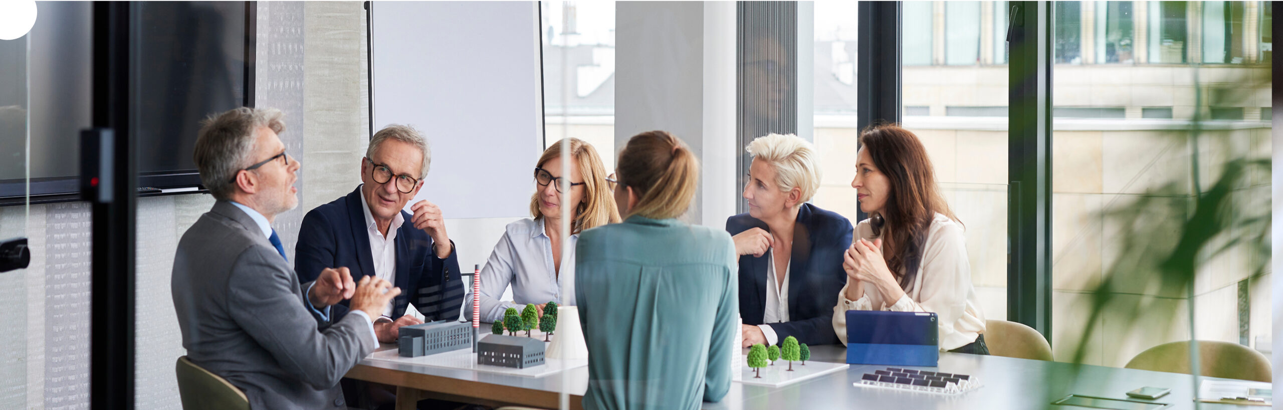 A group of six professionals, three men and three women, sit around a meeting table with architectural models, engaged in discussion in a bright modern office with large windows.