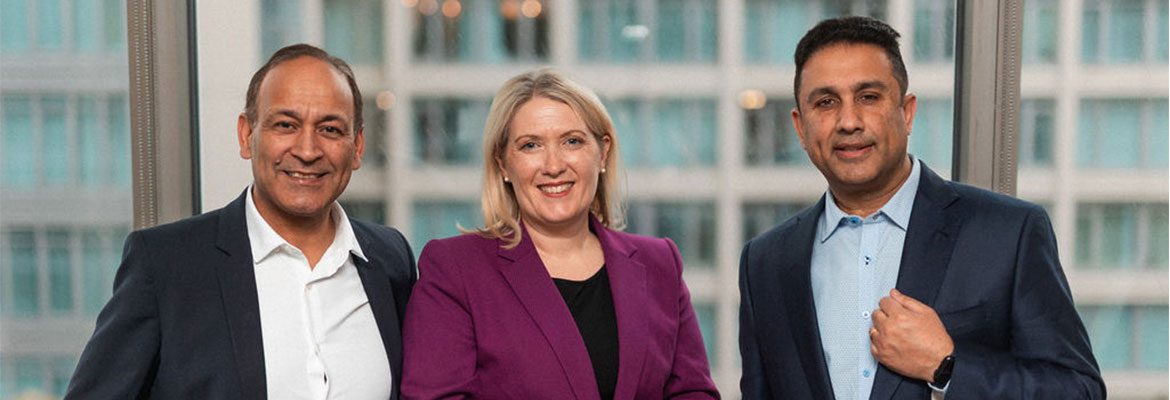 Three people in business attire are standing together and smiling in front of a large window with an office building in the background.