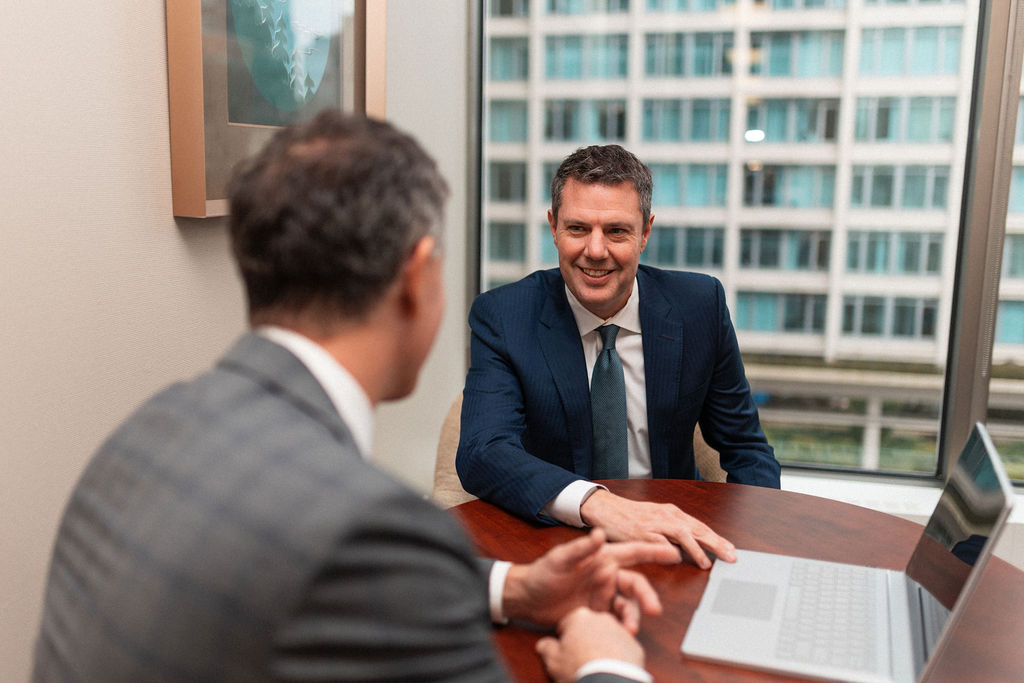 Two men in suits sit at a table in a modern office, engaged in conversation. One man faces the camera, smiling, while the other’s back is to the camera. A laptop and large window with city views are visible.